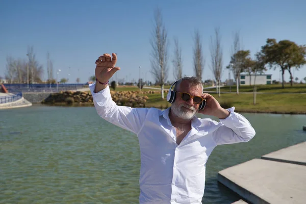 Olgun, sakallı, güneş gözlüklü, beyaz gömlekli, beyaz kulaklıklı, göl kenarında müzik dinlerken mutlu bir şekilde dans eden bir adam. Mutluluk kavramı, neşe, müzik, disko, dans, emeklilik.