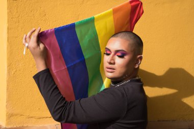 İkili olmayan bir kişinin portresi, genç ve Güney Amerikalı, ağır makyajlı, gözleri kapalı ve elinde bir gay gururu bayrağı var. Kavram kraliçesi, lgbtq +, gurur, nonoş.