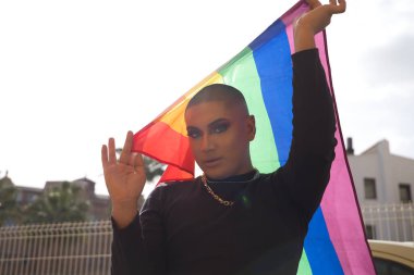 İkili olmayan genç ve Güney Amerikalı 'nın portresi ağır makyaj yapıyor ve arkasında güçlü bir ışık tutan bir gey gururu bayrağı taşıyor. Kavram kraliçesi, lgbtq +, gurur, nonoş.