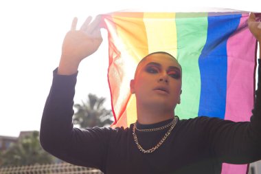 İkili olmayan genç ve Güney Amerikalı 'nın portresi ağır makyaj yapıyor ve arkasında güçlü bir ışık tutan bir gey gururu bayrağı taşıyor. Kavram kraliçesi, lgbtq +, gurur, nonoş.
