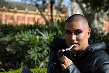 İkili olmayanların portresi, genç ve Güney Amerikalı, ağır makyajlı, dişleriyle ısıran güneş gözlüğü tapınağı ve bitki örtüsüyle çevrili. Kavram kraliçesi, lgbtq +, gurur, nonoş.