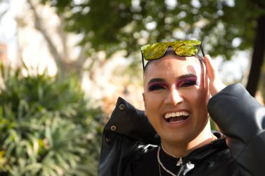 İkili olmayan birinin portresi, genç ve Güney Amerikalı, makyajlı, mutlu bir şekilde gülümseyen ve kafasında aynalı güneş gözlüğü olan ve yeşilliklerle çevrili. Kavram kraliçesi, lgbtq +, gurur, nonoş.