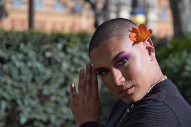 İkili olmayan genç ve Güney Amerikalı 'nın portresi, ağır makyajlı, kulağına güzel bir çiçek konmuş ve yeşillikle çevrili. Kavram kraliçesi, lgbtq +, gurur, nonoş, çiçekler.