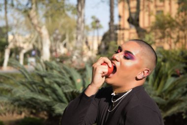 İkili olmayan bir kişinin portresi, genç ve Güney Amerikalı, ağır makyajlı, elinde tuttuğu ve bitkilerle çevrili kırmızı bir elmayı ısırıyor. Konsept kraliçesi, lgbtq +, gurur, ibne, günah.