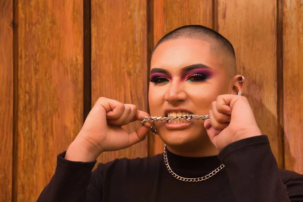 İkili olmayan bir kişinin portresi, genç ve Güney Amerikalı, arka planda tahta bir duvar bulunan dişli metal bir zinciri ısırıyor. Kavram kraliçesi, lgbtq +, gurur, nonoş.