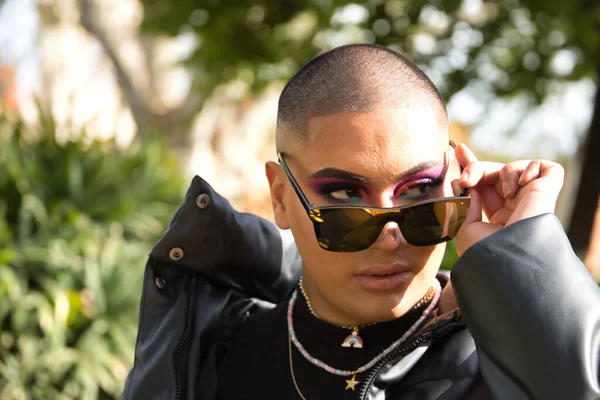 İkili olmayan genç ve Güney Amerikalı 'nın portresi, ağır makyajlı, aynalı güneş gözlüklerine bakıyor ve yeşilliklerle çevrili. Kavram kraliçesi, lgbtq +, gurur, nonoş.