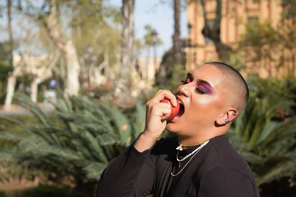 İkili olmayan bir kişinin portresi, genç ve Güney Amerikalı, ağır makyajlı, elinde tuttuğu ve bitkilerle çevrili kırmızı bir elmayı ısırıyor. Konsept kraliçesi, lgbtq +, gurur, ibne, günah.