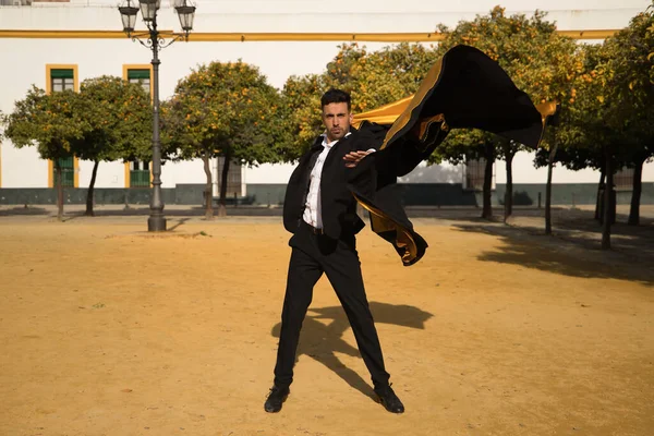 Young Spanish man in black shirt, jacket and pants, with dancing shoes, dancing flamenco with black and gold capote in the street. Typical Spanish concept, art, dance, culture, tradition.