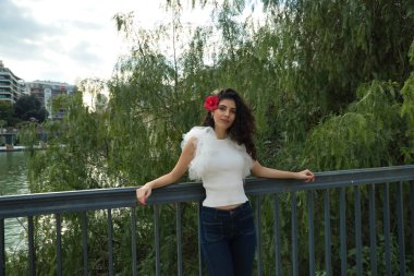 Genç ve güzel bir kadın, esmer, kıvırcık saçlı, kulağında kırmızı bir çiçek, beyaz gömlek ve kot pantolon giyiyor, bir ağacın yanındaki korkuluğa yaslanıyor. Güzellik, moda, çiçekler, doğa kavramları.