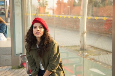 Young and beautiful woman, brunette, with curly hair, with green jacket and red beret, sitting at a stop waiting for the bus. Beauty concept, fashion, trend, public transport, buses.