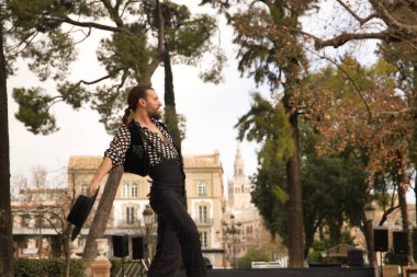 Sakallı, at kuyruklu, siyah tişörtlü, beyaz benekli, siyah pantolonlu ve ceketli, sahnede şapkalı flamenko dansı yapan genç bir adam. Sanat, dans, kültür, gelenek.