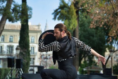 Sakallı, at kuyruklu, siyah tişörtlü, beyaz benekli, siyah pantolonlu ve ceketli, sahnede şapkalı flamenko dansı yapan genç bir adam. Sanat, dans, kültür, gelenek.