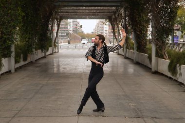 Sakallı, at kuyruklu, siyah tişörtlü, beyaz benekli, siyah pantolonlu ve ceketli, şehrin ortasında flamenko dansı yapan genç bir adam. Sanat, dans, kültür, gelenek.