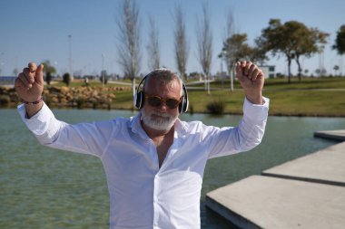 Olgun, sakallı, güneş gözlüklü, beyaz gömlekli, beyaz kulaklıklı, göl kenarında müzik dinlerken mutlu bir şekilde dans eden bir adam. Mutluluk kavramı, neşe, müzik, disko, dans, emeklilik.