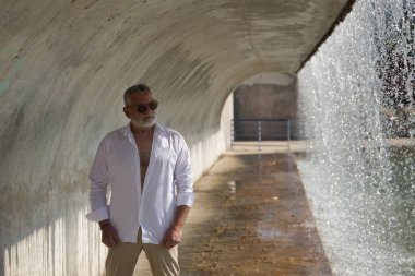 Olgun, gri saçlı, sakallı, güneş gözlüklü, beyaz gömlekli, açık kahverengi pantolonlu, şelalenin yanındaki beton tünelin altında. Olgun bir adam, zengin bir baba, yaşam tarzı, oyuncak ayı.