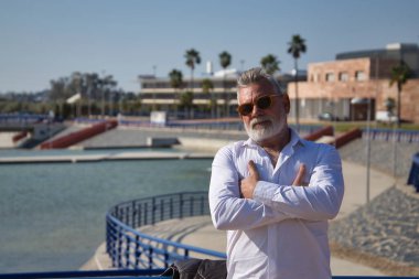 Mature, gray-haired man with beard, sunglasses, white shirt and arms crossed, leaning on a railing in the open air. Concept grandfather, sugar daddy, lifestyle, mature man.