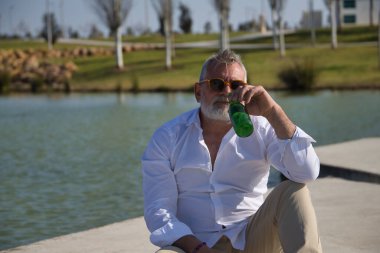 Olgun, sakallı, güneş gözlüklü, beyaz gömlekli, açık kahverengi pantolonlu, ağzında bir şişe birayla gölün yanında oturan bir adam. Olgun erkek kavramı, alkol, içki, tatil, sorunlar.