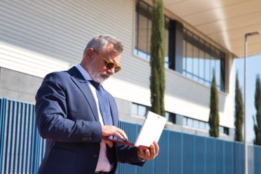 Olgun bir adam, profesör, gri saçlı, sakallı, güneş gözlüklü, ceketli ve kravatlı üniversitenin çıkışındaki dizüstü bilgisayardan e-postaları kontrol eden. Rektör konsepti, iş adamı, iş adamı, uygulamalar, dizüstü bilgisayar.