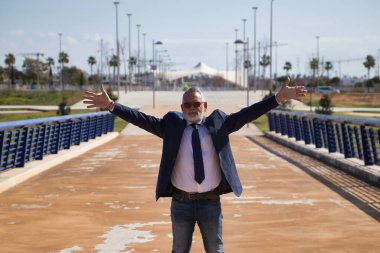 Olgun bir adam, yönetici, gri saçlı, sakallı, güneş gözlüklü, ceketli ve kravatlı, kolları açık, mutlu, çünkü işler onun için iyi gidiyor. Kavram yöneticisi, işadamı, iş adamı, istihdam, zafer.