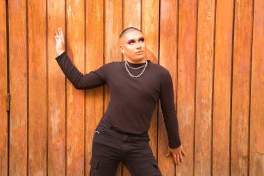 İkili olmayan, genç ve Güney Amerikalı, aşırı makyajlı ve tamamen siyah giyinmiş, arka planda tahta bir duvarla gökyüzüne bakıyor. Kavram kraliçesi, lgbtq +, gurur, nonoş.