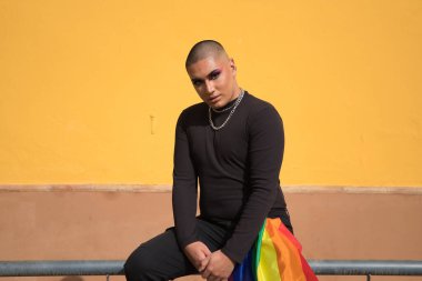 İkili olmayan, genç Güney Amerikalı, ağır makyajlı, pantolon cebinde bir gay gururu bayrağı taşıyan bir korkulukta oturuyor. Kavram kraliçesi, lgbtq +, gurur, nonoş.