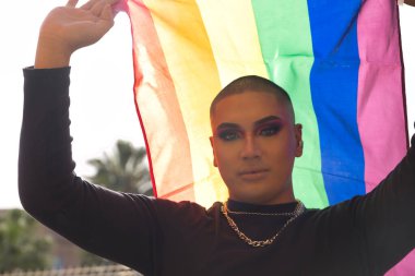 İkili olmayan genç ve Güney Amerikalı 'nın portresi ağır makyaj yapıyor ve arkasında güçlü bir ışık tutan bir gey gururu bayrağı taşıyor. Kavram kraliçesi, lgbtq +, gurur, nonoş.