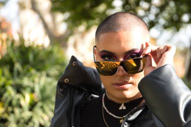 İkili olmayan genç ve Güney Amerikalı 'nın portresi, ağır makyajlı, aynalı güneş gözlüklerine bakıyor ve yeşilliklerle çevrili. Kavram kraliçesi, lgbtq +, gurur, nonoş.