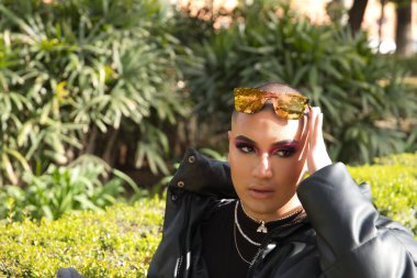 İkili olmayan genç ve Güney Amerikalı birinin portresi. Makyajlı ve kafasında aynalı güneş gözlüğü var. Etrafı yeşillikle çevrili. Kavram kraliçesi, lgbtq +, gurur, nonoş.