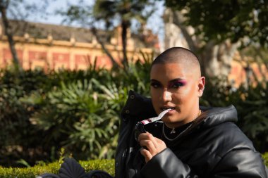 İkili olmayanların portresi, genç ve Güney Amerikalı, ağır makyajlı, dişleriyle ısıran güneş gözlüğü tapınağı ve bitki örtüsüyle çevrili. Kavram kraliçesi, lgbtq +, gurur, nonoş.