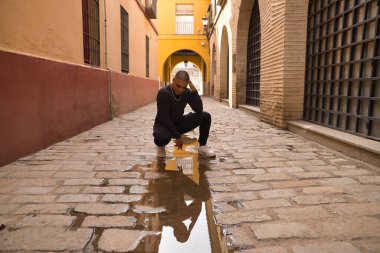 İkili olmayan, genç ve Güney Amerikalı, sokağın ortasında ağır makyajlı, elleri başının üstünde, su birikintisindeki yansımasına endişeyle bakıyor. Kavram kraliçesi, lgbtq +, gurur, nonoş.