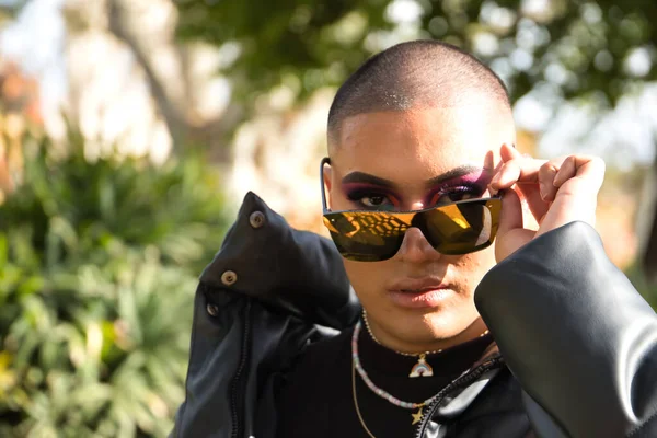 İkili olmayan genç ve Güney Amerikalı 'nın portresi, ağır makyajlı, aynalı güneş gözlüklerine bakıyor ve yeşilliklerle çevrili. Kavram kraliçesi, lgbtq +, gurur, nonoş.