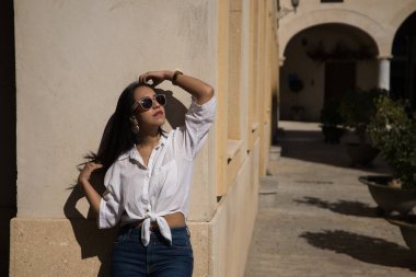 Beyaz gömlekli, kot pantolonlu ve güneş gözlüklü güzel bir Güney Amerikalı kadının portresi Avrupa 'da tatilde bir duvarda güneşleniyor, saçlarına dokunuyor. Güzellik, moda, tatil, seyahat kavramı.