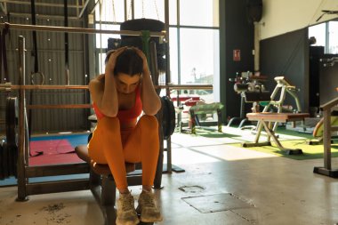 Spor salonundaki bankta oturan genç ve güzel bir kadın elleri başının üstünde, yorgun ve depresif. Depresyon, yorgunluk, stres, spor, yalnızlık, aşırı eğitim.