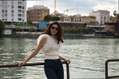 Genç ve güzel bir kadın, esmer, kıvırcık saçlı, beyaz gömlekli, kot pantolonlu ve güneş gözlüklü, nehir kenarındaki korkuluklara yaslanmış. Güzelliği, modayı, modeli, trendi, seyahati kavra.