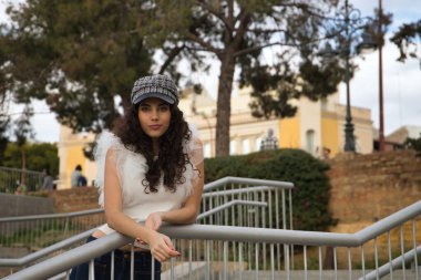 Genç ve güzel bir kadın, esmer, kıvırcık saçlı, beyaz gömlekli, kot pantolonlu ve şapkalı, korkuluğa yaslanmış. Güzellik, moda, model, trend, sükunet kavramı.