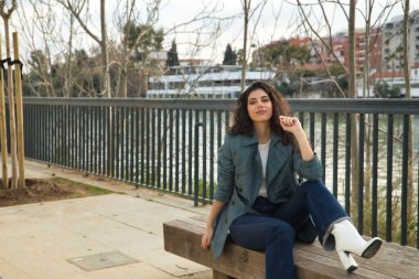 Genç ve güzel bir kadın, esmer, kıvırcık saçlı, ceket giyen, kot giyen, beyaz çizmeli, ahşap bir bankta oturan düşünceli bakışlı. Güzellik, moda, moda, model, düşünce, sonbahar.