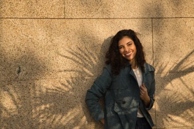 Genç ve güzel bir kadının portresi, esmer, kıvırcık saçlı, ceketli ve güzel bir gülümsemeli, palmiye yapraklarının gölgesinde duvara yaslanmış. Güzellik, moda, sonbahar, gülümseme kavramı.