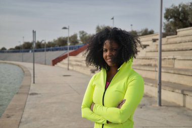 Afro saçlı, spor giyimli, floresan sarı kapüşonlu ve taytlı, kollarını kavuşturmuş, gülümseyen Afro-Amerikalı bir kadın. Fitness konsepti, spor, sokak, şehir.