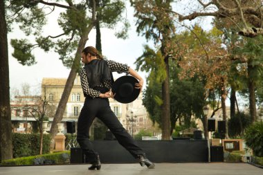 Sakallı, at kuyruklu, siyah tişörtlü, beyaz benekli, siyah pantolonlu ve ceketli, sahnede şapkalı flamenko dansı yapan genç bir adam. Sanat, dans, kültür, gelenek.