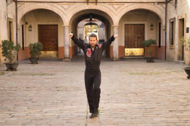 Sakallı, at kuyruklu, siyah şeffaf gömlekli, siyah puantiyeli, kırmızı güllü, siyah pantolonlu ve ceketli, şehirde flamenko dansı yapan genç bir adam. Sanat, dans, kültür, gelenek.