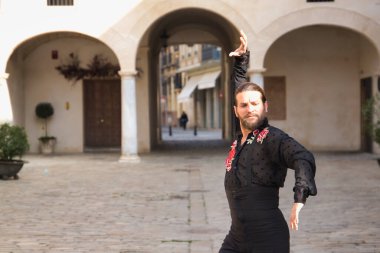 Sakallı, at kuyruklu, siyah şeffaf gömlekli, siyah puantiyeli, kırmızı güllü, siyah pantolonlu ve ceketli, şehirde flamenko dansı yapan genç bir adam. Sanat, dans, kültür, gelenek.