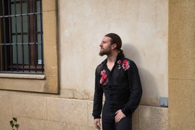 Sakallı, at kuyruklu, flamenko dansçısı, siyah şeffaf gömlekli, puantiyeli, kırmızı güllü, siyah pantolonlu ve ceketli, duvara yaslanmış genç bir adam. Sanat, dans, kültür kavramı.