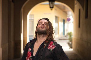 Sakallı, uzun saçlı, siyah şeffaf gömlekli, siyah puantiyeli, kırmızı güllü, siyah pantolonlu ve ceketli, şehirde flamenko dansı yapan genç bir adam. Sanat, dans, kültür, gelenek.