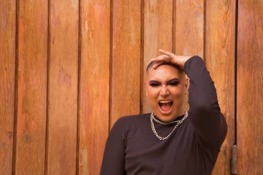 İkili olmayan birinin portresi, genç ve Güney Amerikalı, ağır makyajlı ve arka planda ahşap bir duvarla çığlık çığlığa bağırıyor. Kavram kraliçesi, lgbtq +, gurur, nonoş.