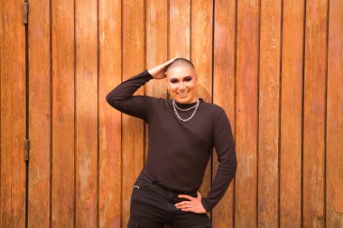 İkili olmayan, genç ve Güney Amerikalı, aşırı makyajlı ve tamamen siyah giyinmiş, arka planda tahta bir duvarla gülümsüyor. Kavram kraliçesi, lgbtq +, gurur, nonoş.