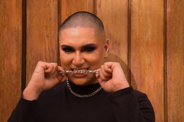 İkili olmayan bir kişinin portresi, genç ve Güney Amerikalı, arka planda tahta bir duvar bulunan dişli metal bir zinciri ısırıyor. Kavram kraliçesi, lgbtq +, gurur, nonoş.