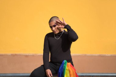 İkili olmayan, genç Güney Amerikalı, ağır makyajlı, pantolon cebinde bir gay gururu bayrağı taşıyan bir korkulukta oturuyor. Kavram kraliçesi, lgbtq +, gurur, nonoş.