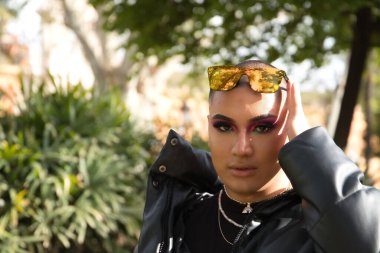 İkili olmayan genç ve Güney Amerikalı birinin portresi. Makyajlı ve kafasında aynalı güneş gözlüğü var. Etrafı yeşillikle çevrili. Kavram kraliçesi, lgbtq +, gurur, nonoş.