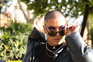 İkili olmayan genç ve Güney Amerikalı 'nın portresi, ağır makyajlı, aynalı güneş gözlüklerine bakıyor ve yeşilliklerle çevrili. Kavram kraliçesi, lgbtq +, gurur, nonoş.