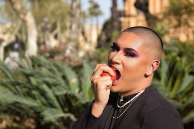 İkili olmayan bir kişinin portresi, genç ve Güney Amerikalı, ağır makyajlı, elinde tuttuğu ve bitkilerle çevrili kırmızı bir elmayı ısırıyor. Konsept kraliçesi, lgbtq +, gurur, ibne, günah.